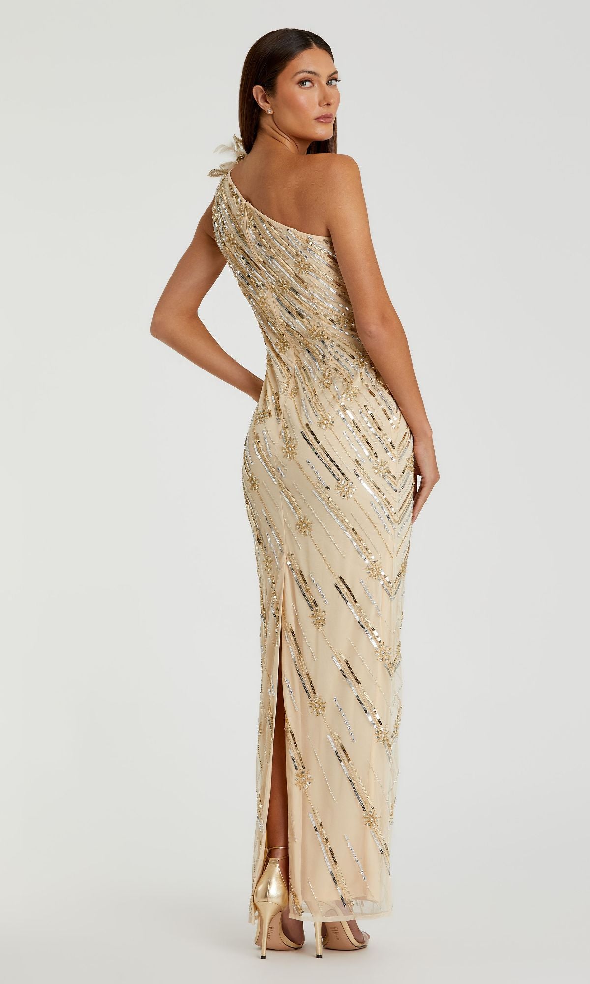 A woman in a Mac Duggal 93739 long gold sequin formal dress with a one-shoulder design and back slit, paired with gold high heels, stands against a plain light background.